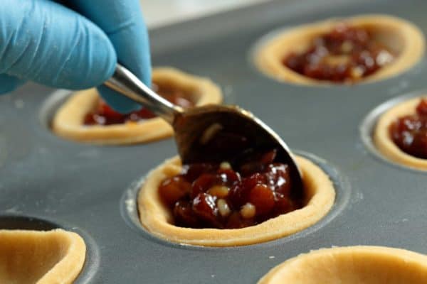 Person putting mince pie filling onto pastry base in a metal baking tray with a spoon